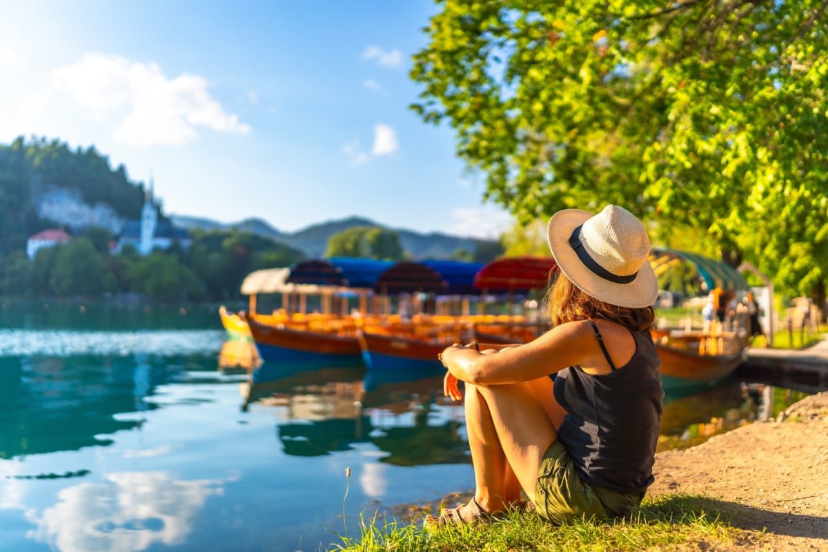 Young woman in summer attire and hat relaxing by lake bled in slovenia, enjoying views of traditional pletna boats and the church